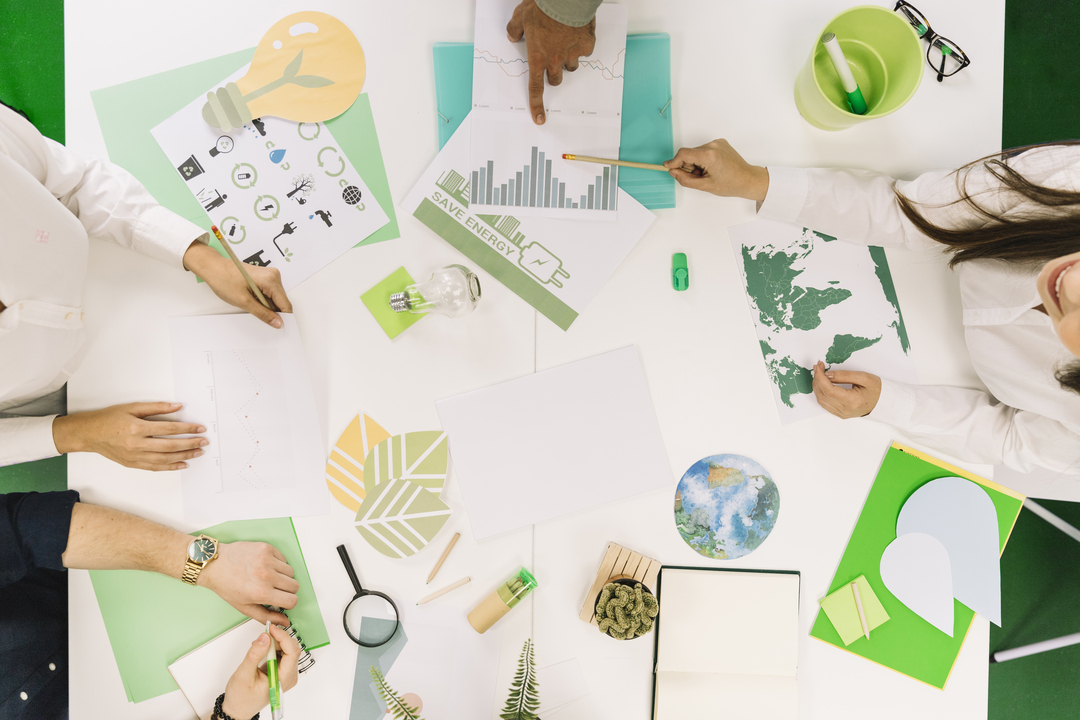 Sustentabilidade na indústria: foto panorâmica de uma mesa com papéis brancos e verdes com as mãos de pessoas.