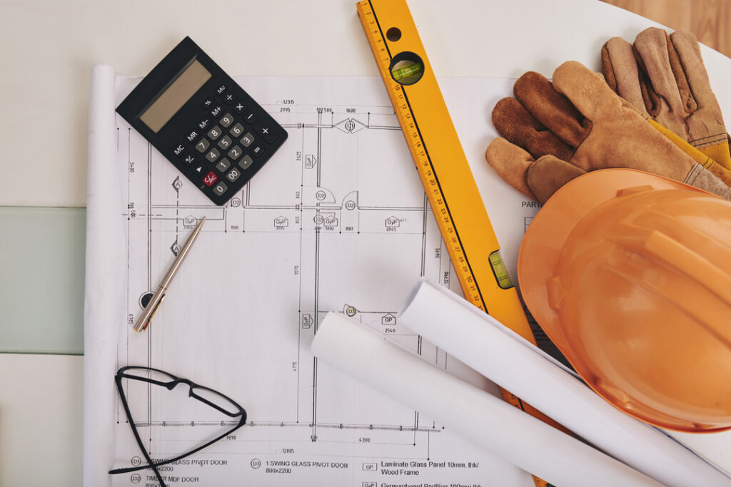 Na foto, vemos uma calculadora, um óculos, luvas, capacete e v[arios projetos sob uma mesa, assim como uma régua ára medição das peças personalizadas.